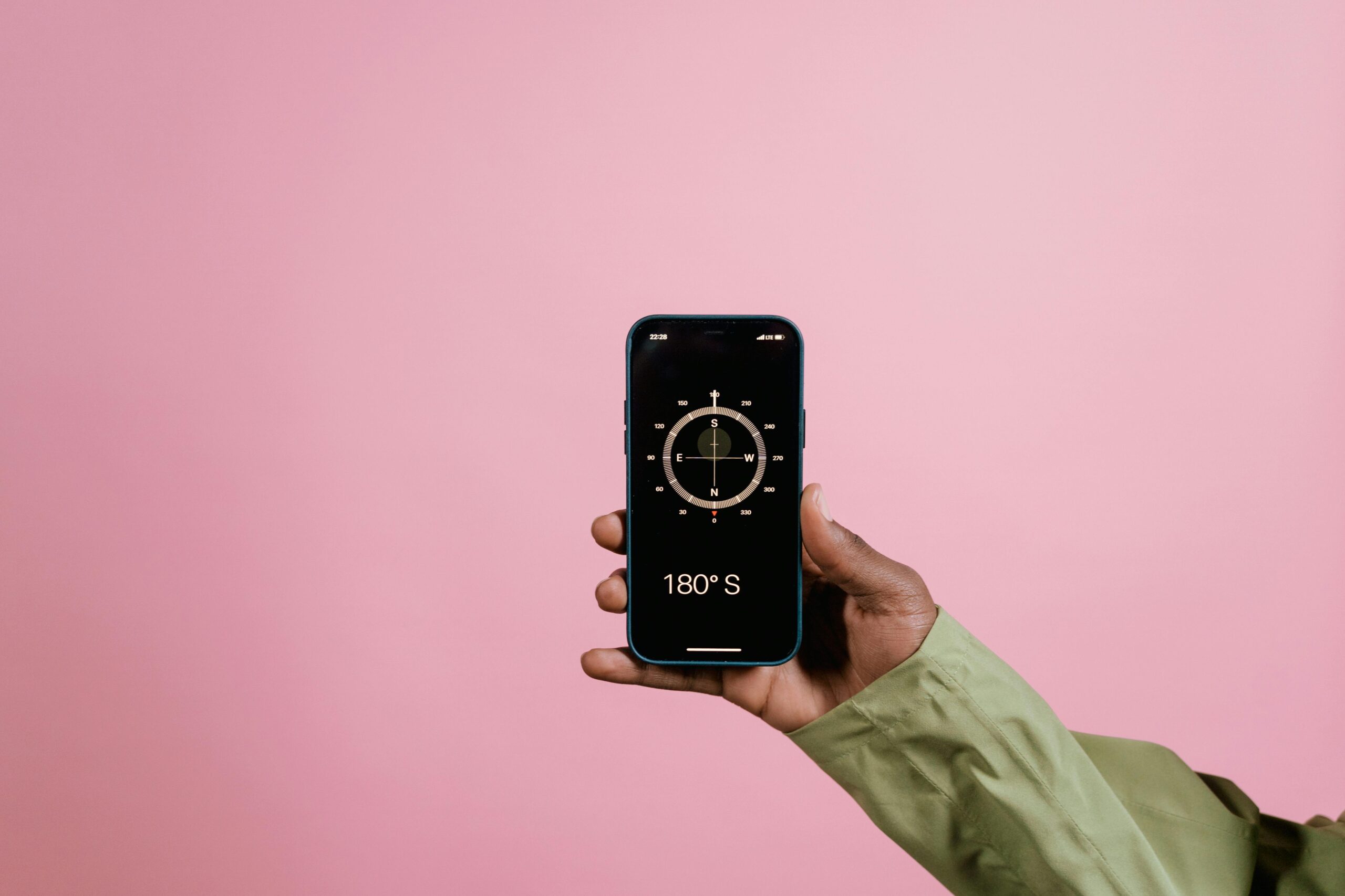 Close-up of a hand holding a smartphone displaying a compass app against a pink background.